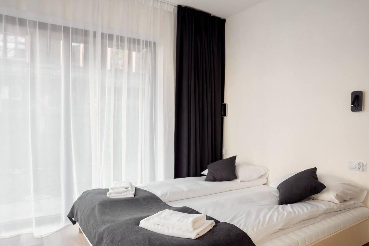 Une chambre d’hôtel avec deux lits, une grande fenêtre avec des rideaux légers, un rideau noir et une lampe fixée au mur.