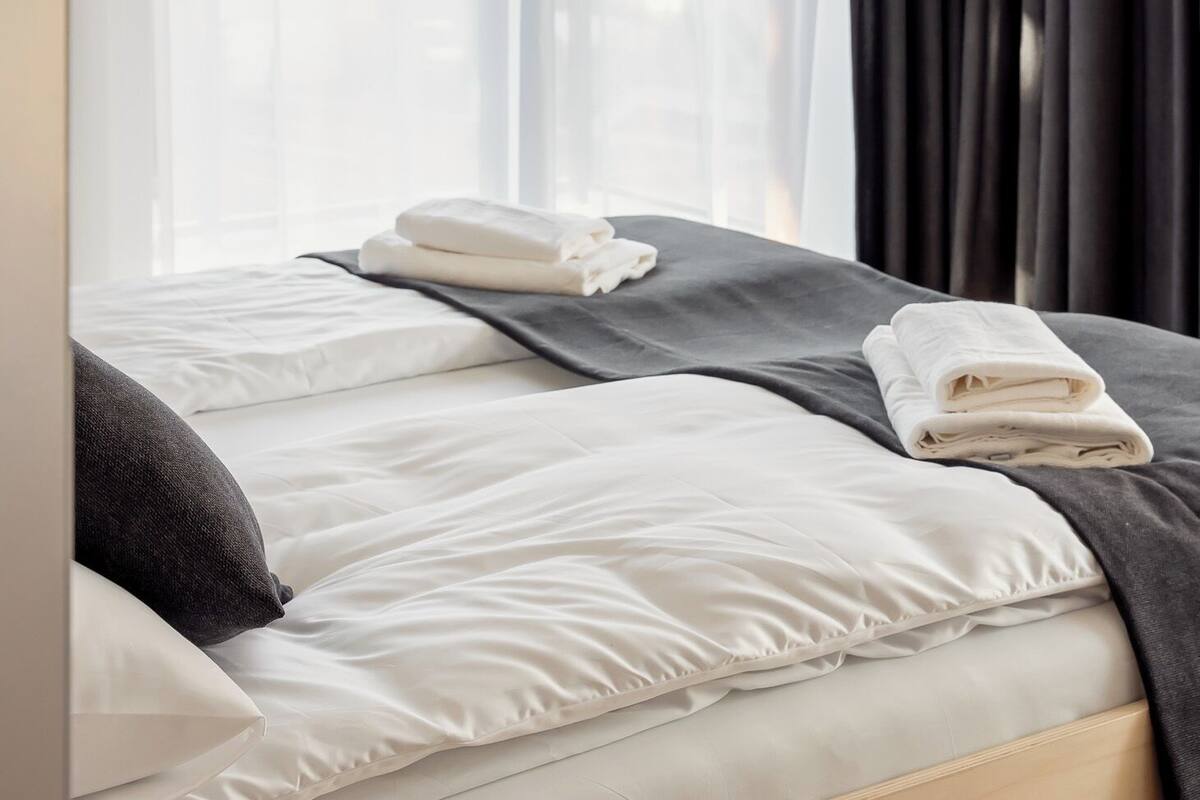 Une chambre d’hôtel avec un lit, du linge de lit blanc, une couverture gris foncé et une fenêtre avec des rideaux légers.