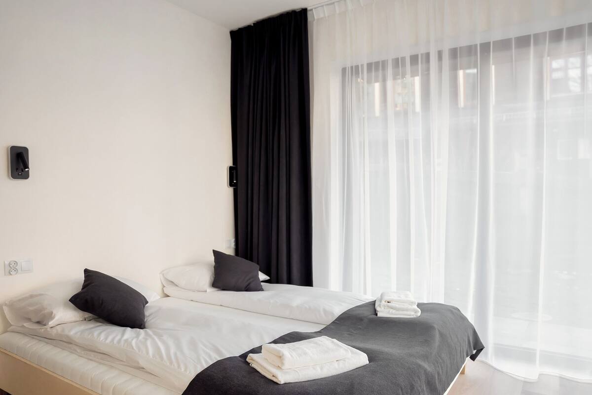 Une chambre d’hôtel avec deux lits, une grande fenêtre avec des rideaux légers et un rideau noir à la fenêtre.