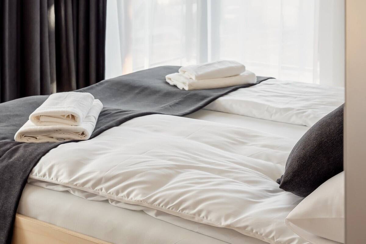 Une chambre d’hôtel avec un lit, du linge de lit blanc, une couverture gris foncé et une fenêtre avec des rideaux légers.