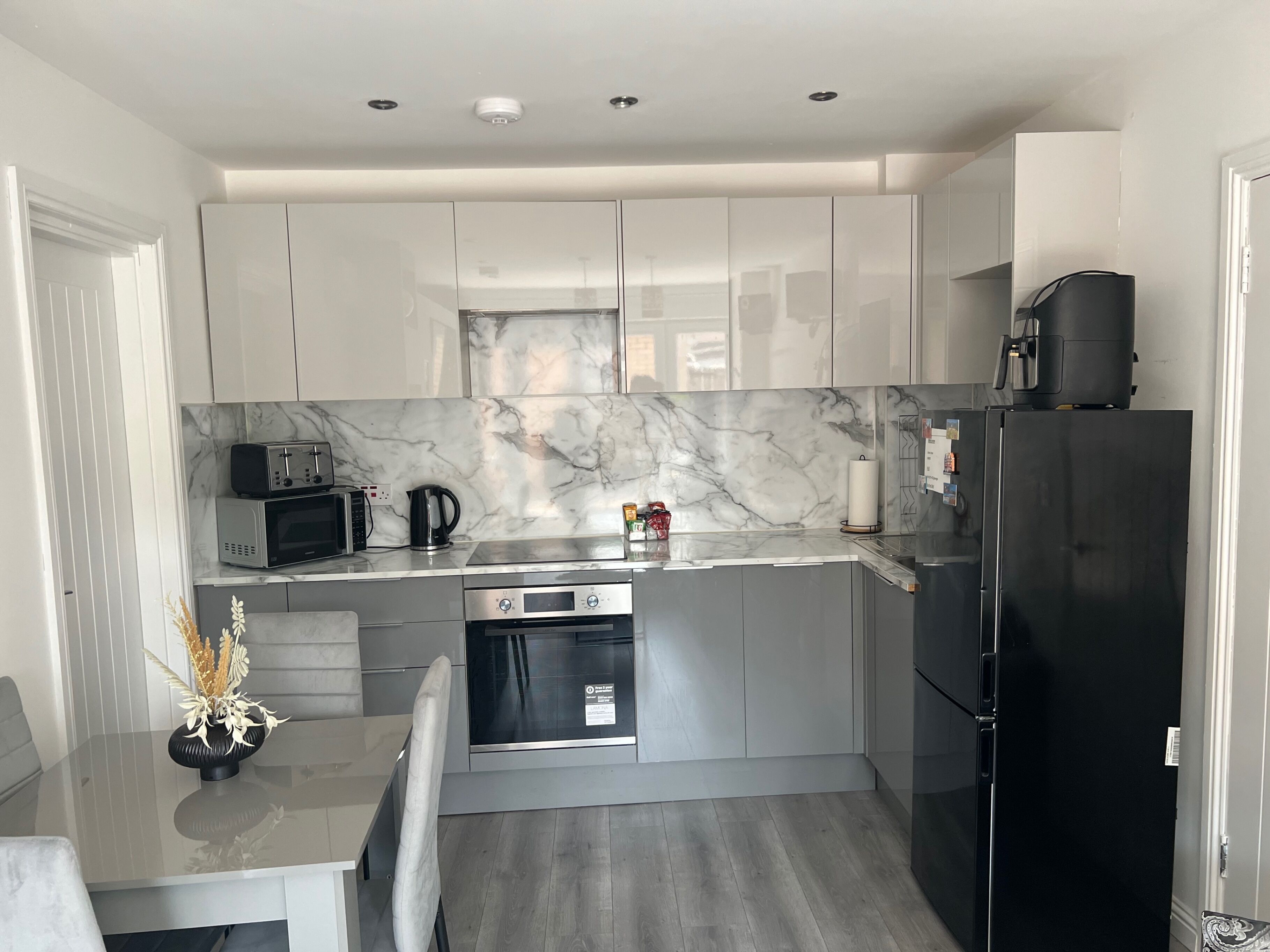 Una cocina moderna con armarios blancos, salpicadero de mármol, refrigerador negro y una zona de comedor con mesa y sillas.