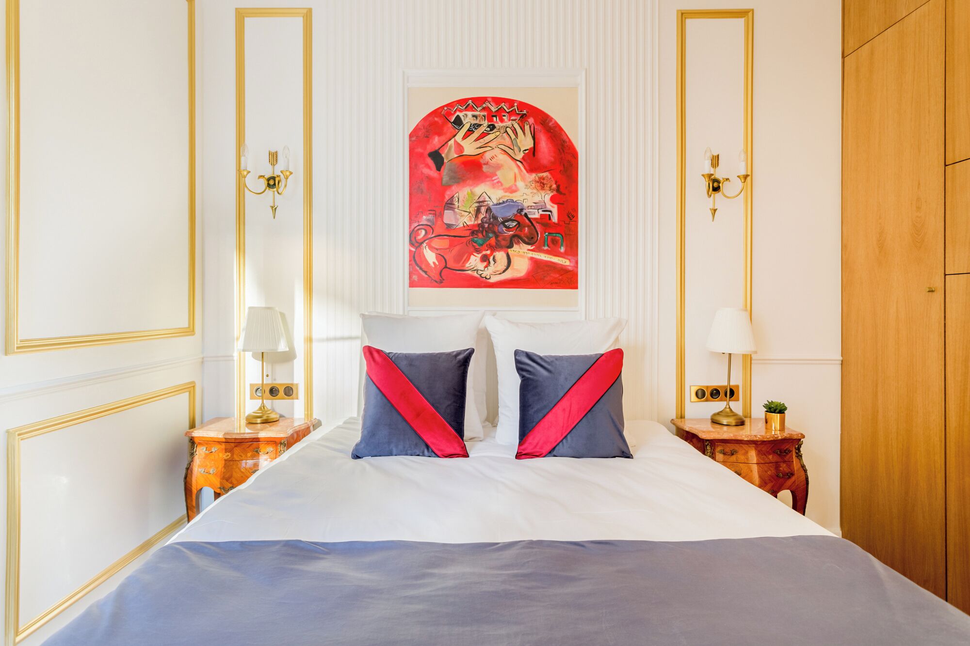 A hotel room with a large bed, two bedside tables with lamps, a framed artwork above the bed, and a wooden wardrobe.