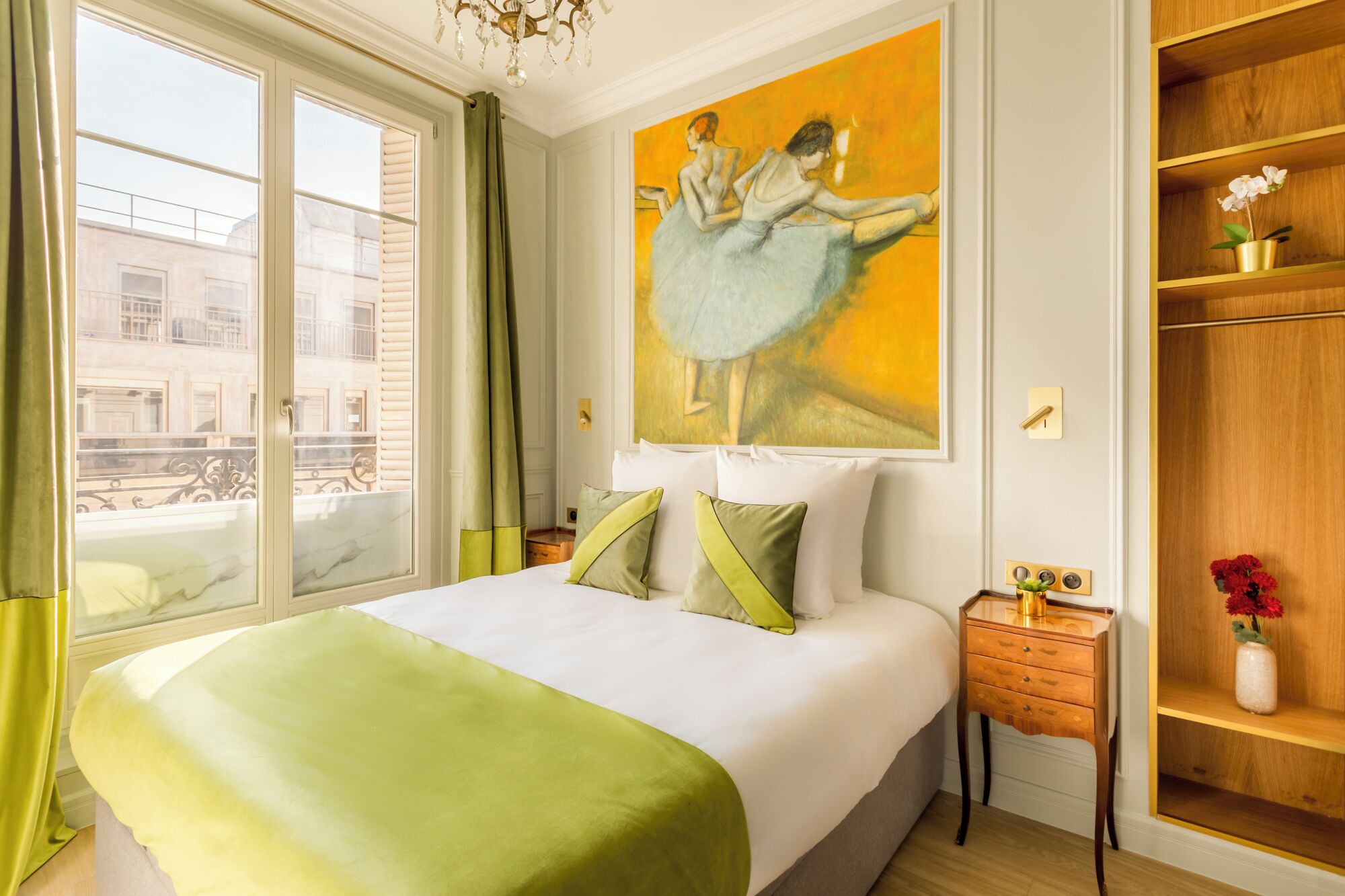 A hotel room with a large bed, a painting of ballerinas, a wooden wardrobe, and a view of the city through the window.