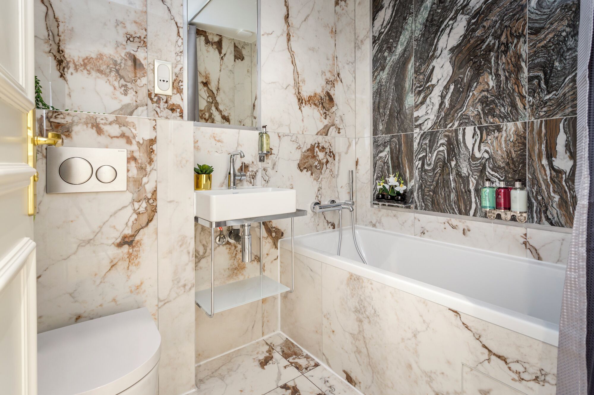 Un baño con pared de mármol, lavamanos, inodoro, bañera y espejo.