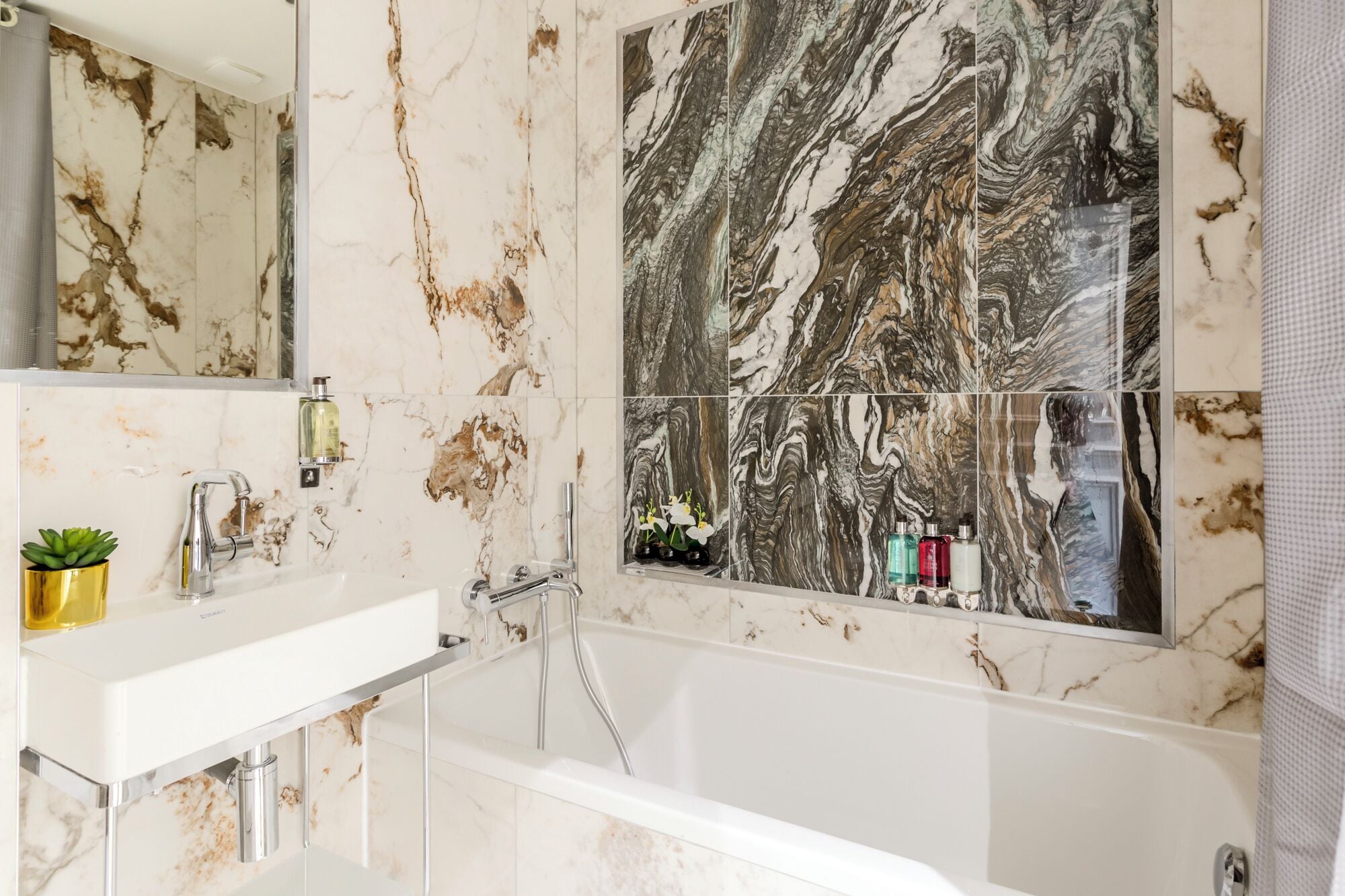 A bathroom with a marble wall, a white sink, a bathtub, and a mirror.