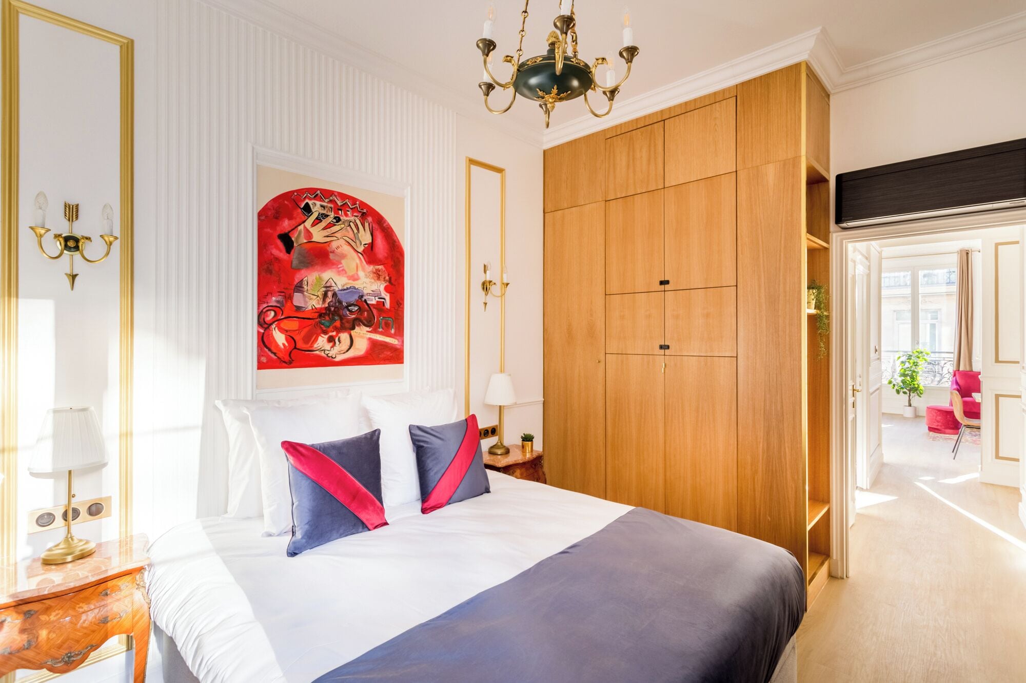 A hotel room with a large bed, a wooden wardrobe, a chandelier, and a painting on the wall.