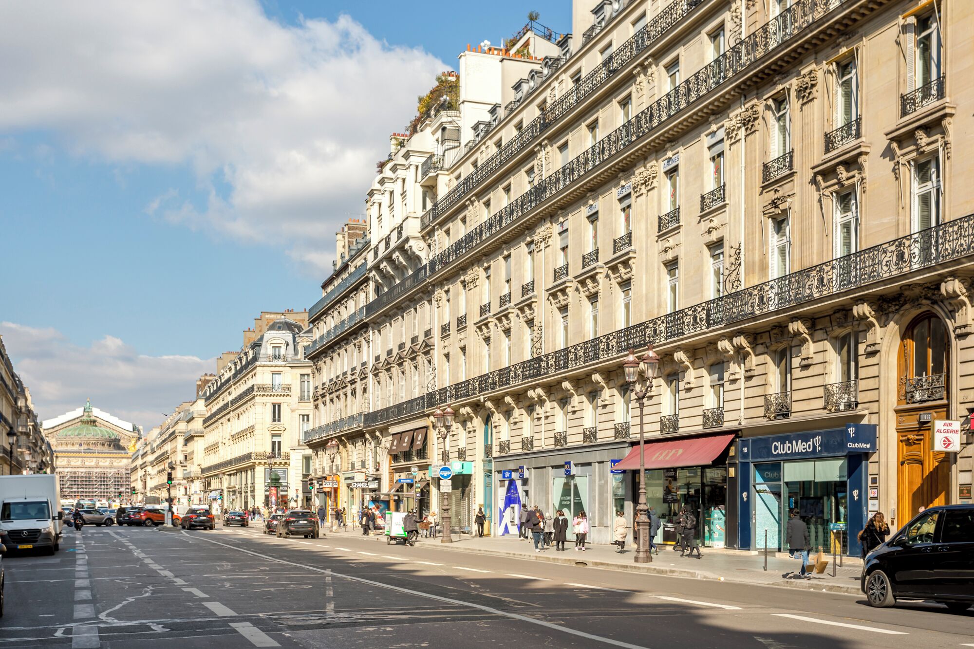 Une vue d’une rue de Paris, montrant un immeuble à l’architecture classique, avec des balcons richement décorés et une rangée de boutiques au rez-de-chaussée.