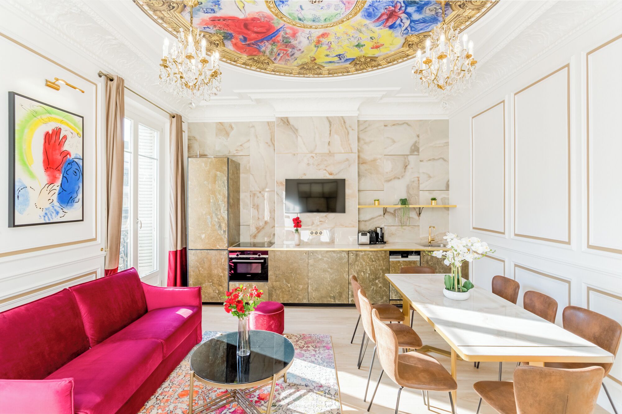Een ruime woonkamer met een rode bank, een eethoek met een witte tafel en bruine stoelen, een keuken met marmeren wanden en een kleurrijk plafond.