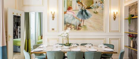 A dining room with a long table set for six, green upholstered chairs, and a large painting of ballerinas on the wall.