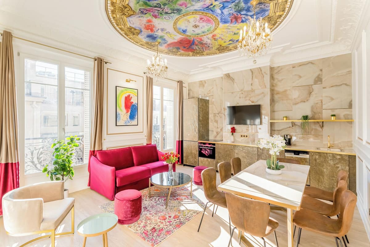 A spacious living room with a red sofa, a dining table with chairs, a chandelier, and a large window with curtains.