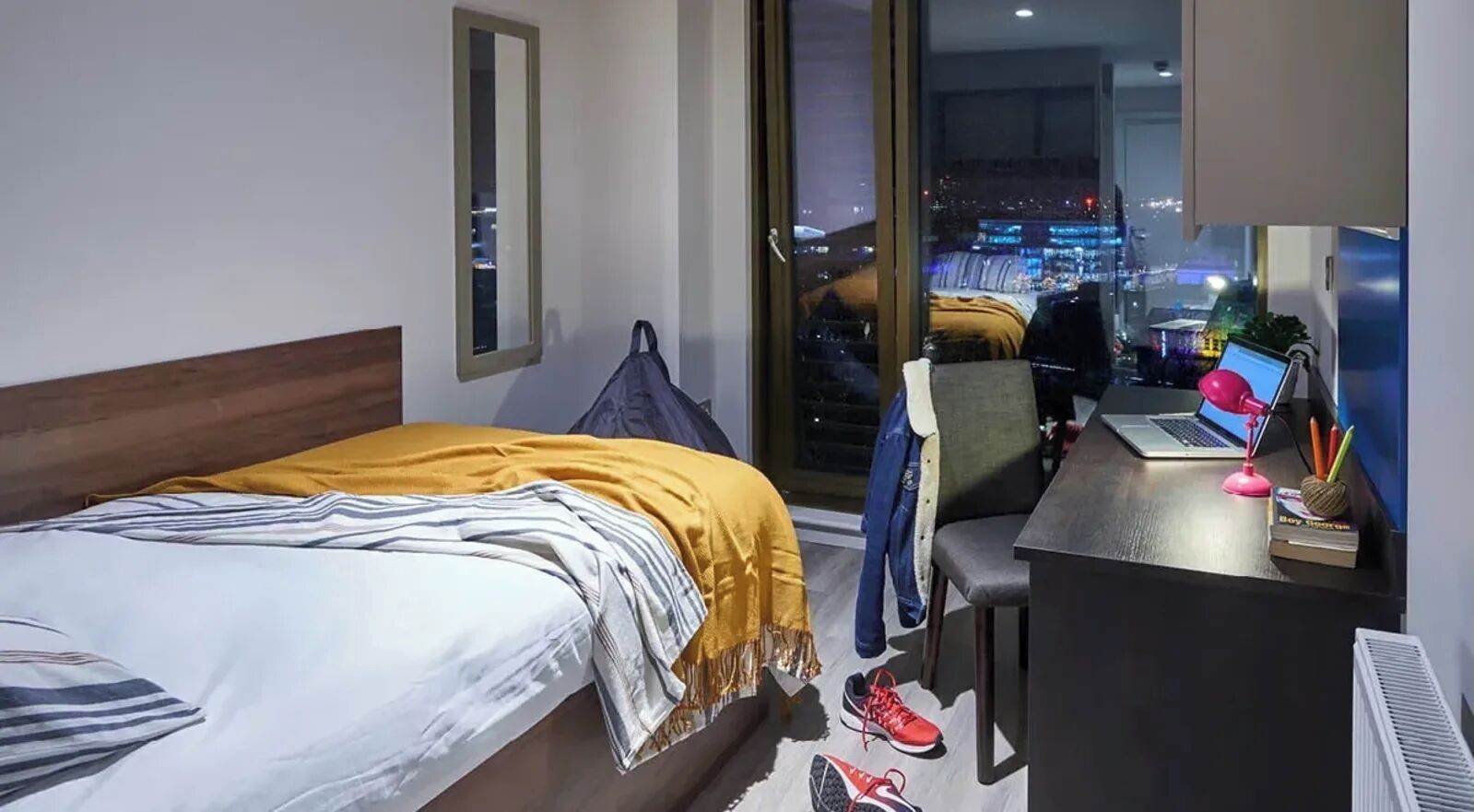 Hotelkamer met een bed, een bureau met een laptop, een stoel en uitzicht op een stadsgezicht door een groot raam.