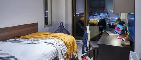 Hotelkamer met een bed, een bureau met een laptop, een stoel en uitzicht op een stadsgezicht door een groot raam.