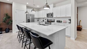 Cozinha moderna com armários brancos, uma ilha grande com banquetas, eletrodomésticos de aço inoxidável e iluminação pendente.