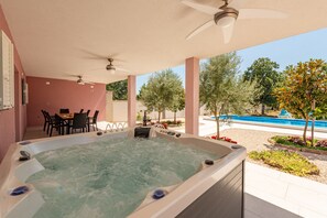 Un jacuzzi con una mesa y sillas cerca, un ventilador de techo y vistas a una piscina y árboles.