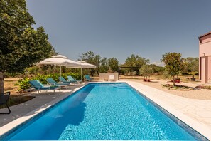 Una piscina rectangular con agua cristalina azul, rodeada por una solana con baldosas. Hay sillas de descanso y sombrillas, y se ve un edificio al fondo.