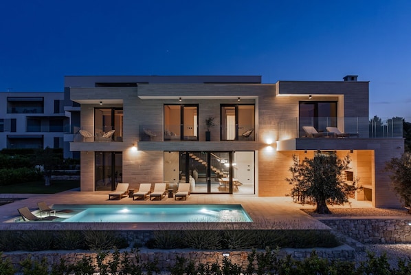 A modern two-story house with a swimming pool, lounge chairs, and a well-lit exterior.