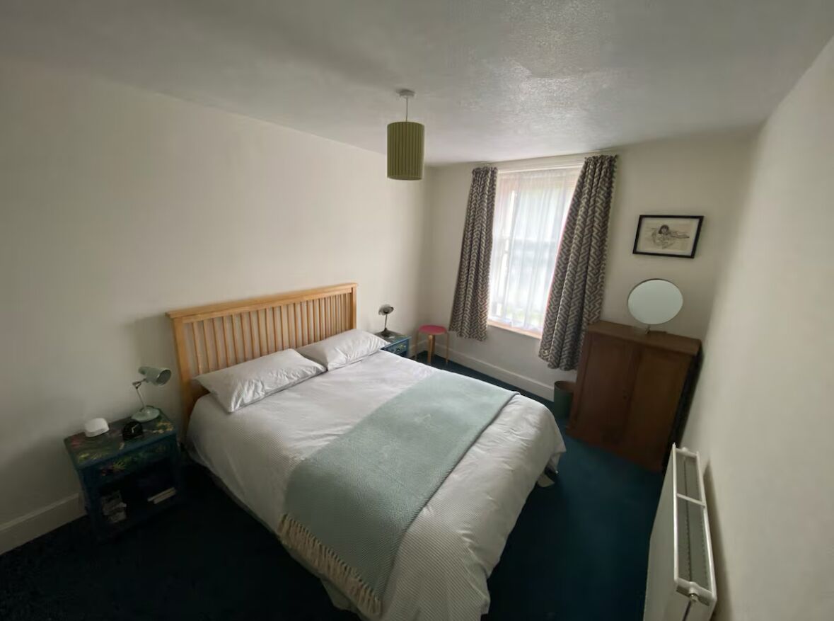 A bedroom with a bed, bedside table, lamp, window with curtains, and a picture on the wall.