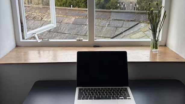 Un ordinateur portable est posé sur un bureau près d’une fenêtre, d’où l’on aperçoit une église et des arbres.