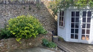 Un mur de pierres avec un arbuste, une terrasse en bois et un bâtiment blanc doté de fenêtres.