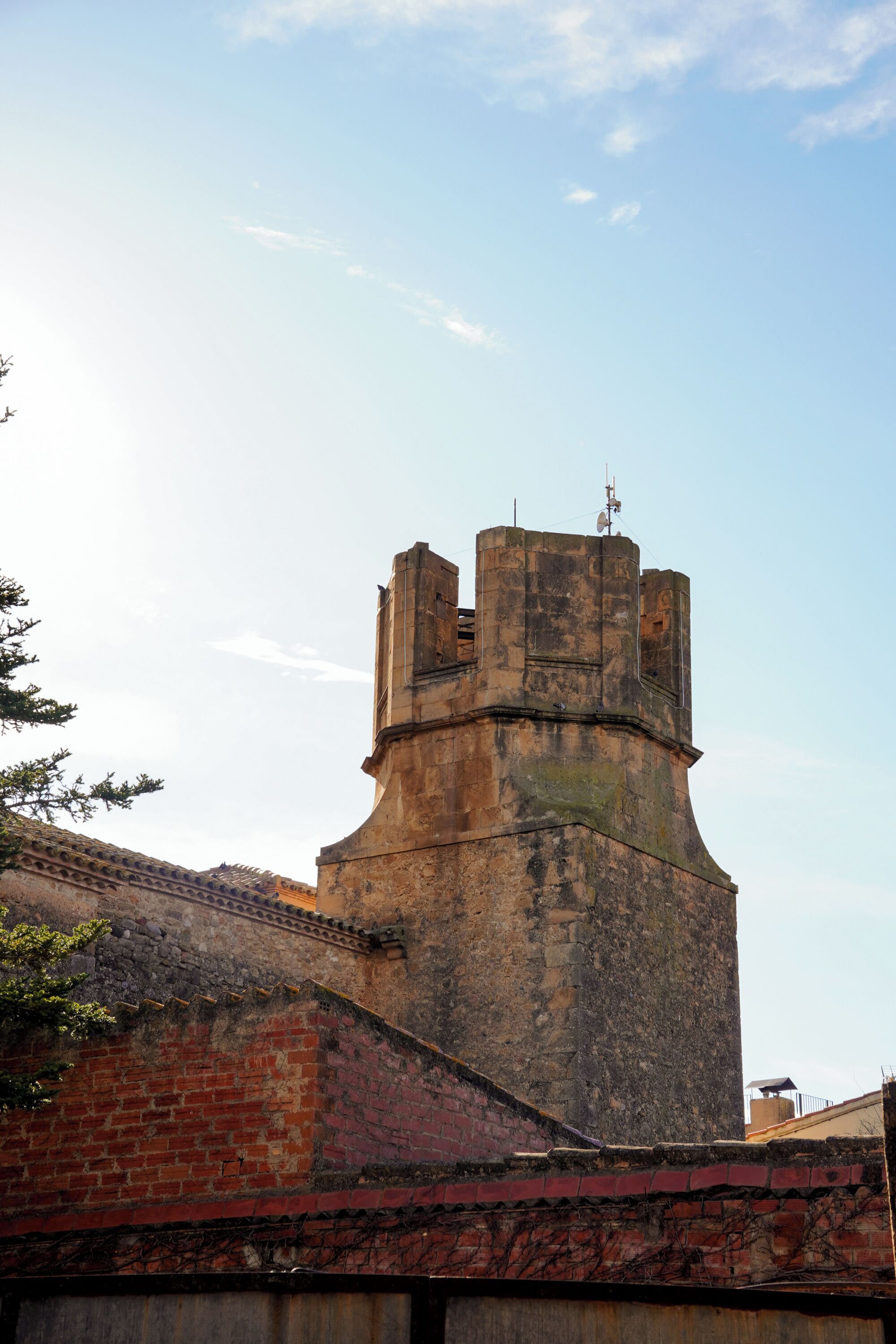 Une tour de pierre historique, surmontée d’une girouette, un mur de briques et un ciel dégagé.