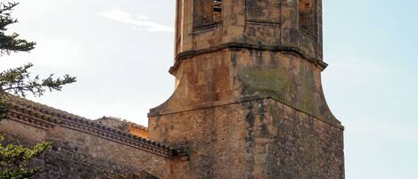 Une tour de pierre historique, surmontée d’une girouette, un mur de briques et un ciel dégagé.