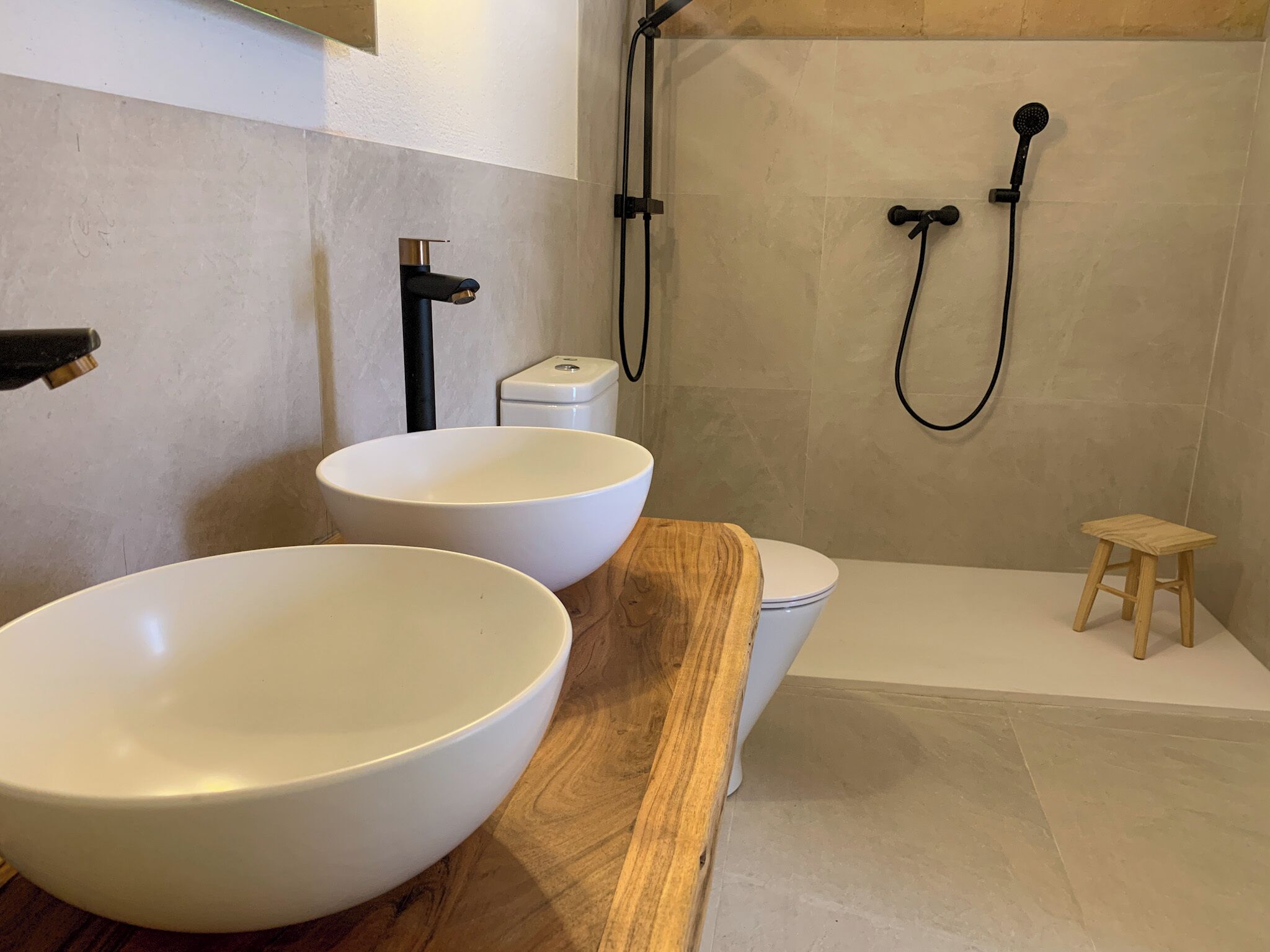 Un baño con dos recipientes blancos grandes sobre una encimera de madera, un inodoro y una zona de ducha con cabezal de ducha manual.