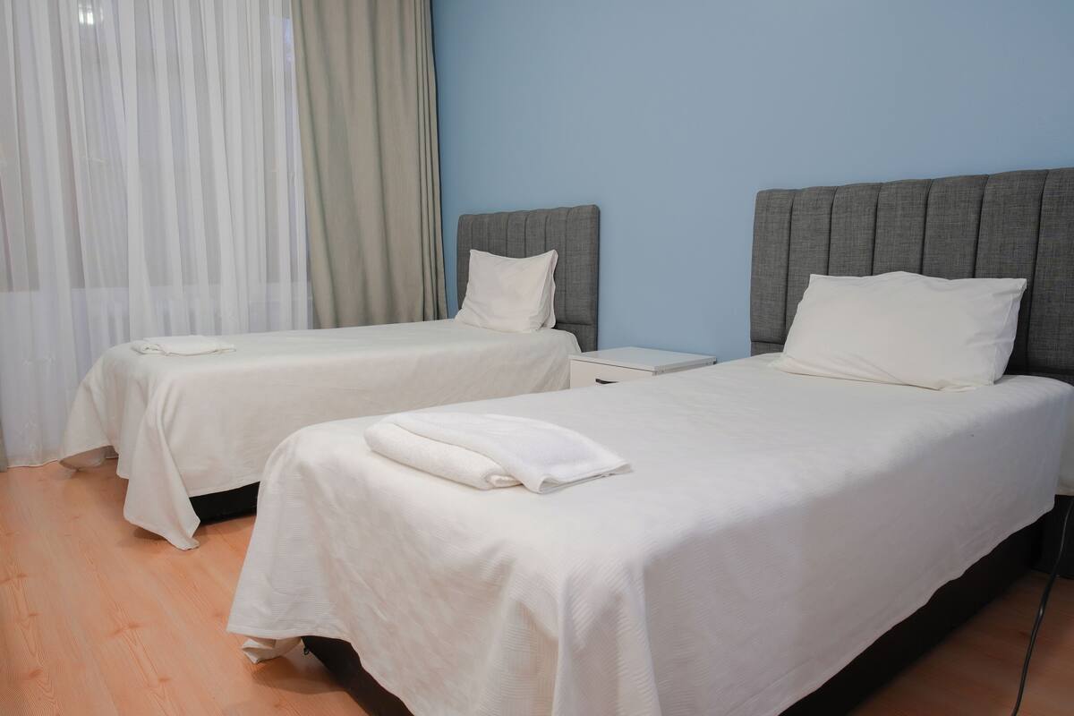 A hotel room with two single beds, a bedside table, a nightstand, and a window with sheer curtains.