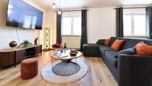 Een moderne woonkamer met een flatscreen televisie, een ronde salontafel, een bank en een groot raam met gordijnen.