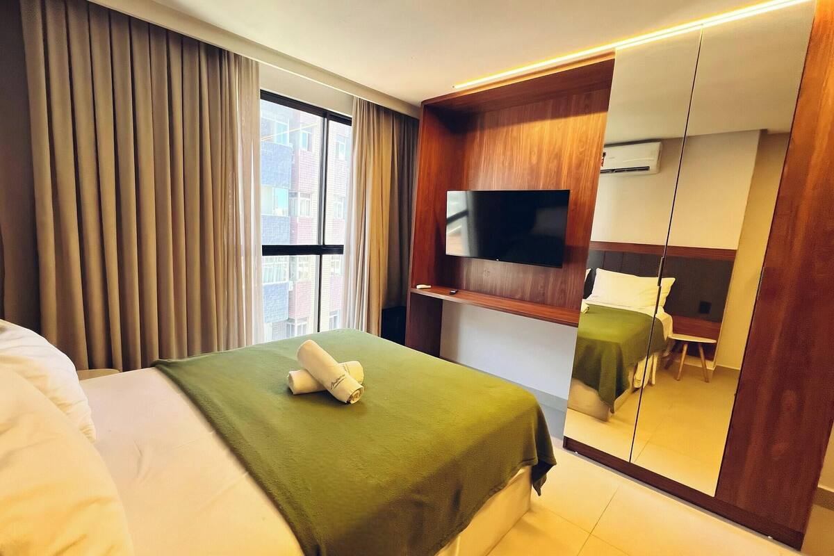 A hotel room with a bed, a TV, a mirror, and a view of buildings outside.