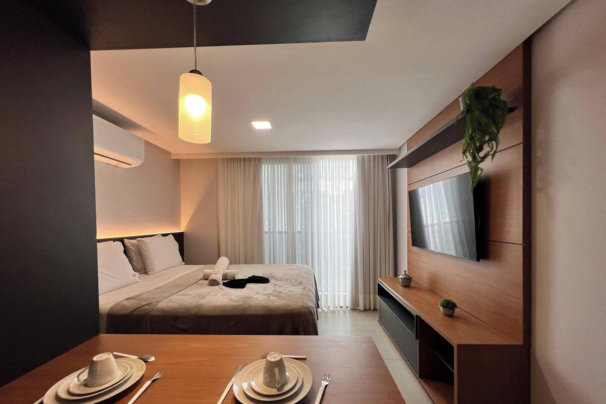 A hotel room with a bed, a TV mounted on the wall, a dining table with plates and cutlery, and a hanging plant.