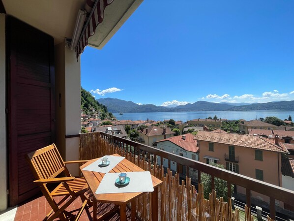 A balcony with two wooden chairs and a table, overlooking a lake with buildings and mountains in the background.