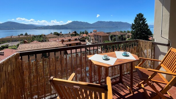 A balcony with wooden chairs and a table, overlooking a lake and mountains.