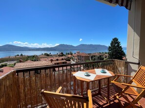 A balcony with wooden chairs and a table, overlooking a lake and mountains.
