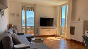 A modern living room with a sofa, a flat-screen TV, and a balcony with a view of the sea and mountains.