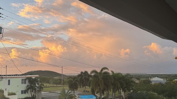A view of a residential area with a swimming pool surrounded by palm trees, a small building, and a hilly landscape in the background. - Sunsets, Breezes & Caribbean Views • Private Puerto Rico Getaway (Salinas)