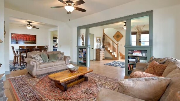 A spacious living room with a beige sofa, a wooden coffee table, and a large rug. There is a staircase leading to an upper level, and a bookshelf filled with books. - Historic Sears Roebuck House (Thomas)