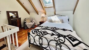 Ein Schlafzimmer mit schrägem Dach, einem Oberlicht, einem Bett mit gemustertem Überwurf, einem Sessel und einem kleinen Tisch mit Lampe.
