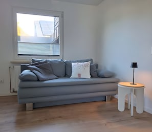 A modern living room with a grey sofa, a small white side table, and a window with blinds.