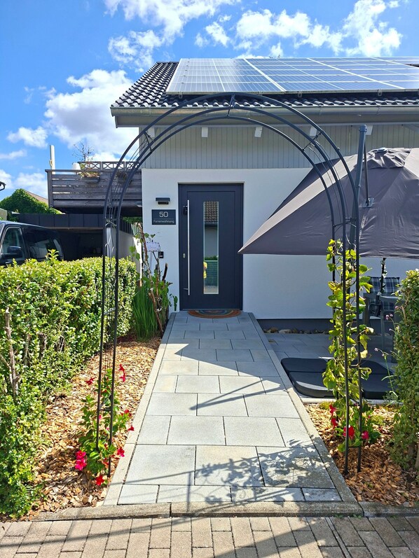 A pathway leading to a building with solar panels on the roof, a door with a number sign, and a covered area with plants.
