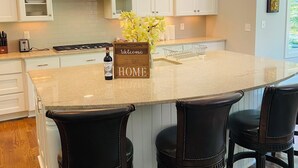 Cocina con armarios blancos, una isla central con taburetes de bar, iluminación colgante y un cartel de bienvenida.