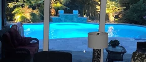 Una habitación con un ventanal que da a una piscina, una silla, una lámpara y una pequeña mesa.