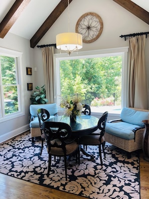A dining area with a round table, chairs, a chandelier, and a view of greenery outside.