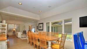 A dining area with a wooden table and chairs, a kitchen in the background, and a large window with a view.