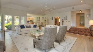 A spacious living room with a white sofa, grey armchairs, and a coffee table. There is a fireplace with a brick surround, and a ceiling fan.