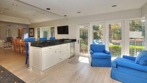 A modern kitchen with white cabinets, a black countertop, and a dining area with wooden chairs. There is a large window providing a view of the outdoors.