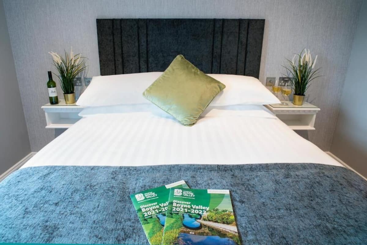 Una cama bien hecha con un almohadón verde, un edredón blanco y una manta azul. Dos mesitas de noche con plantas en macetas y una botella de vino en cada una. Dos folletos están colocados sobre la cama.