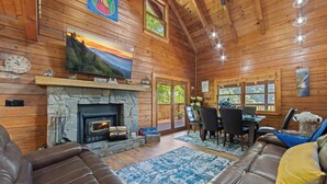 Un intérieur de chalet chaleureux avec une cheminée en pierre, des canapés en cuir, des murs en bois et un coin repas offrant une vue sur l’extérieur.