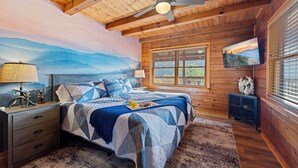 Une chambre dans une cabane en bois, avec un lit, des tables de chevet, un ventilateur de plafond, un tableau, une télévision et une fenêtre avec des stores.