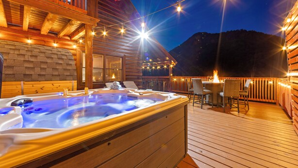 Un bain à remous sur une terrasse en bois, agrémentée de guirlandes lumineuses, avec des murs en bois et une vue sur les montagnes en arrière-plan.