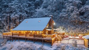 Une cabane en rondins dotée d’une grande terrasse et d’un espace salon extérieur, entourée d’une forêt enneigée.